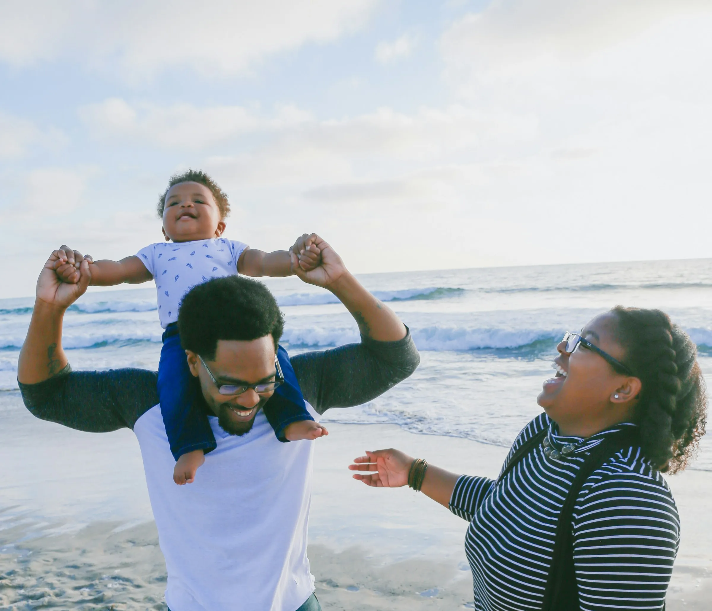 A dad carrying a laughing toddler on his shoulders on the beach beside mom