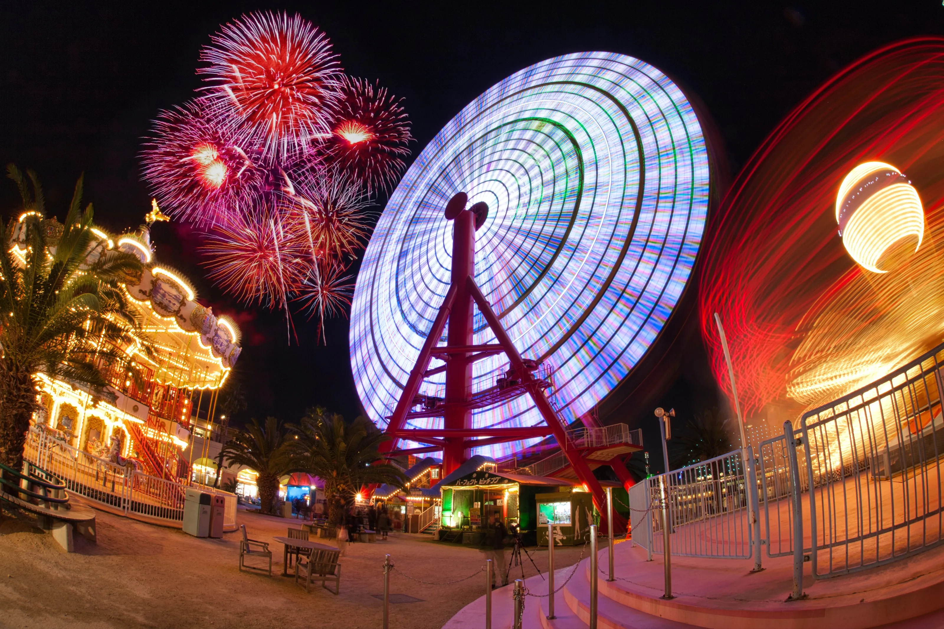 A theme park midway at night with a lit ferris wheel, carousel and fireworks